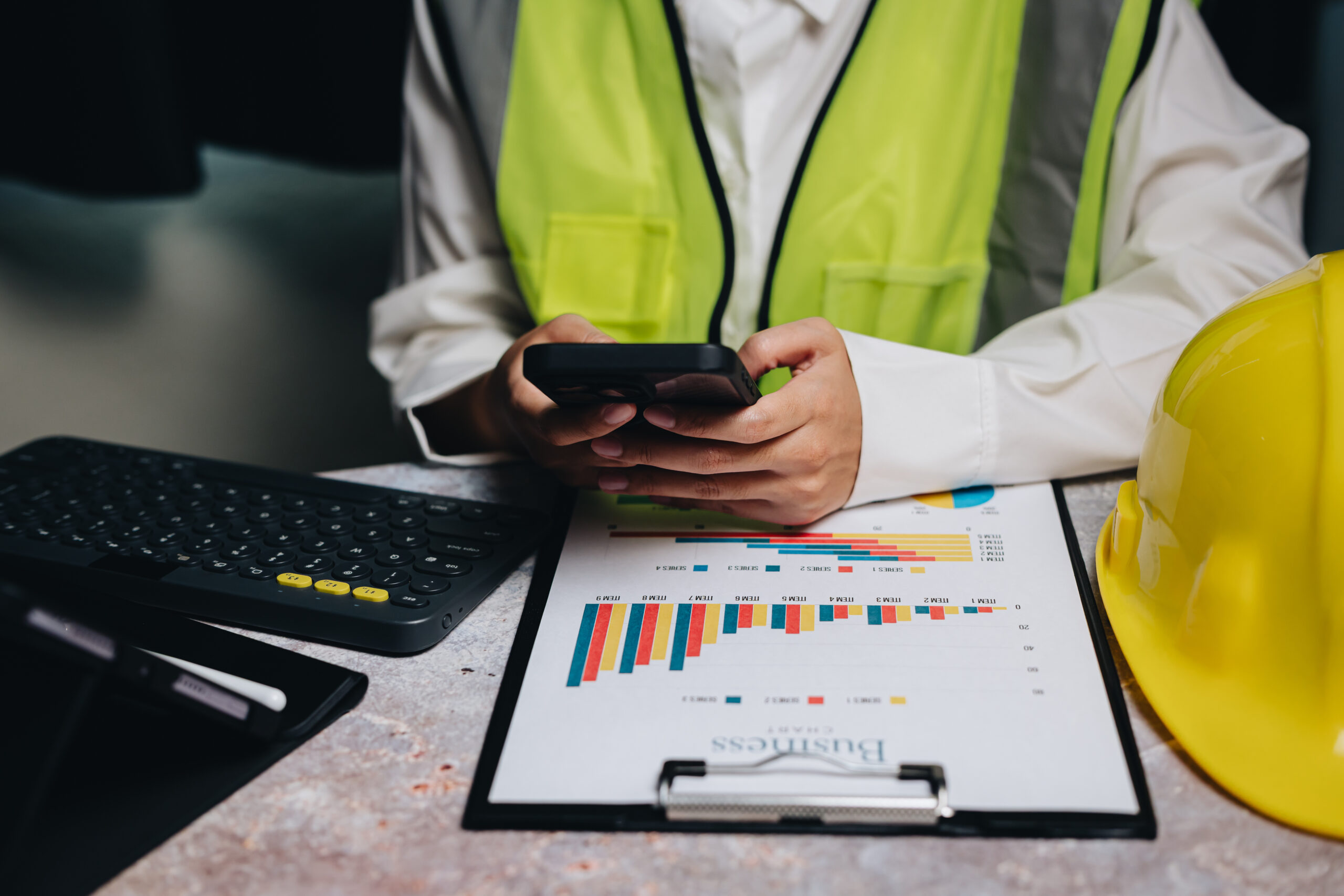 Professional worker in safety vest using smartphone near charts and hard hat in an office setting for business analysis.