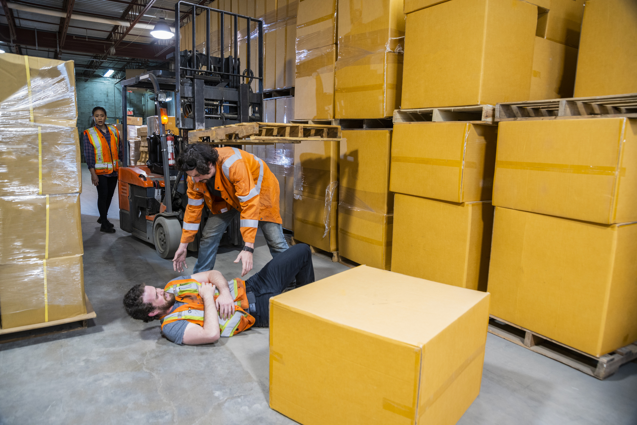 An industrial warehouse workplace safety topic. A worker injured falling or being struck by a forklift. Falls and collisions are major contributors to forklift safety. Coworkers come to his aid.
